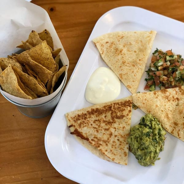 Quesadillas with guacamole, salsa, sour cream, and a bucket of tortilla chips on a wooden table.