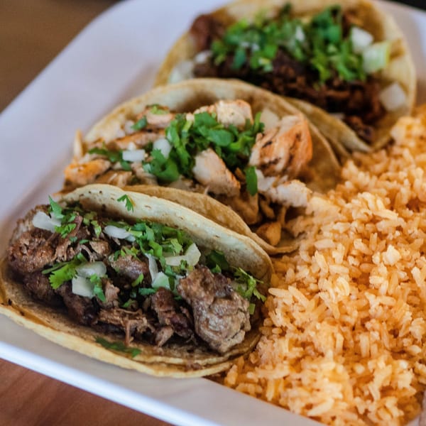 Three Mexican tacos with various fillings, cilantro, and onions, served with a side of rice.
